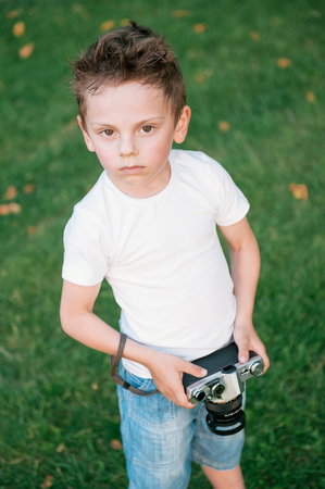 Portrait of beautiful kid wearing shirt and shorts with retro film camera on green grass backdrop in parkの写真素材