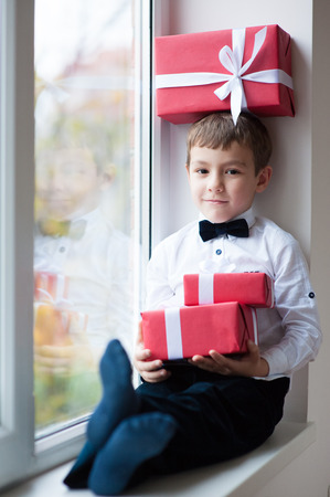beautiful little boy sitting by the window with a gift box on his head and two gift boxes in handsの写真素材
