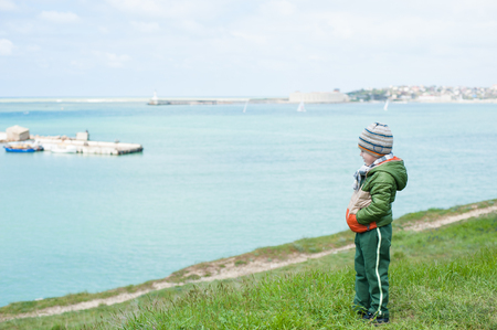 lonely little boy in jacket and hat stands on slope against background of sea in springの写真素材