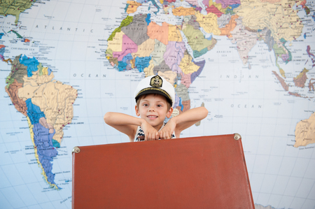 beautiful small boy in the captain's hat raises the suitcase against the background of the world mapの写真素材