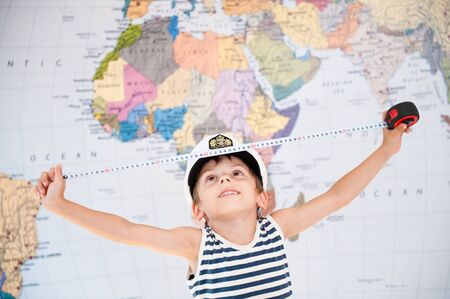 cute little boy in captain hat measures distance on world map with measuring tape