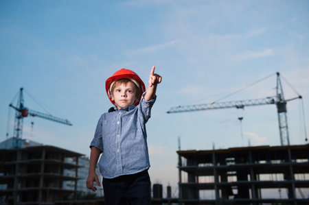 cute child in builder helmet points finger forward standing against silhouettes of cranes and structures of new buildingsの写真素材
