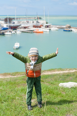 cute small boy dressed in warm jacket scarf and hat jumps on green slope against background of sea harbor with boats in cold springの写真素材