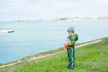 cute pensive little boy in scarf jacket and hat on green grassy slope against sea with floating yachtsの写真素材