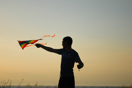 silhouette of active young man controlling colorful kite flying in air at sunsetの写真素材