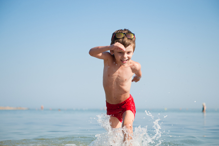 active happy little kid wearing sunglasses and red shorts running in sea water with splashes in summer holidayの写真素材