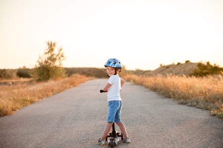 smiling little boy in sport helmet standing on abandoned road with scooter in summer sunsetの写真素材