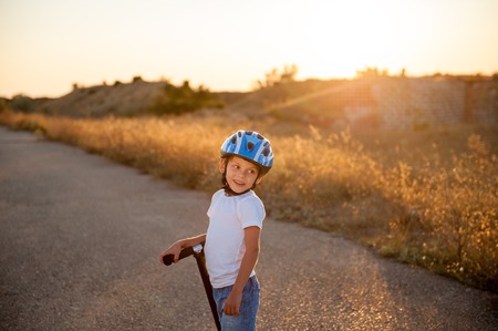 smiling little boy in sport helmet standing on abandoned road with scooter in summer sunsetの写真素材