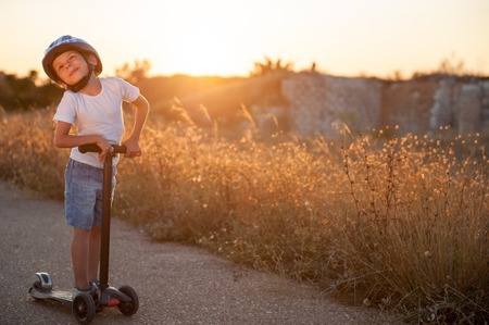happy small boy in sport helmet standing on abandoned road with scooter in warm summer sunsetの写真素材