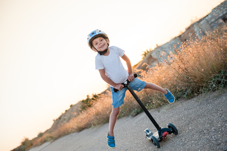 smiling little kid in sport helmet riding on scooter on abandoned road in warm summer sunsetの写真素材