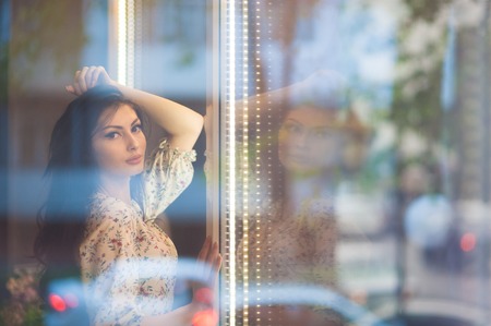 beautiful elegant girl with long hair in summer dress behind glass with reflections of the street in roomの写真素材