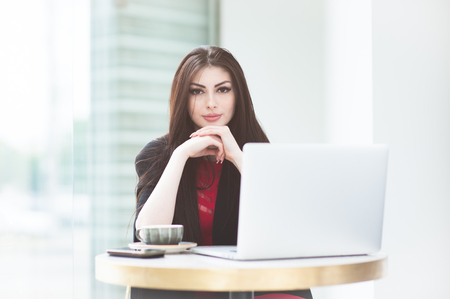 pretty young woman sitting at table with laptop and cup of tea in bright cafe with large window on city streetの写真素材