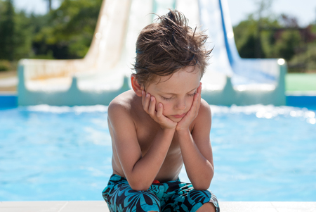 sad little boy sitting near water pool in summer resort water parkの写真素材