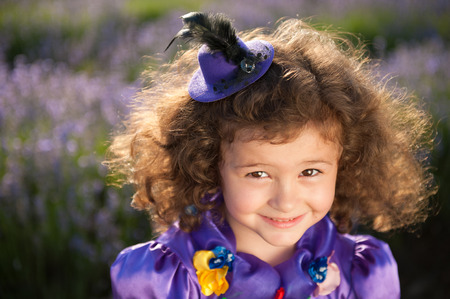 portrait of happy beautiful small girl in little hat and purple dress among flowersの写真素材