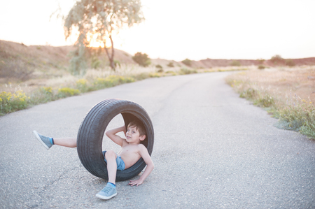 cute little smiling kid in sneakers sitting on highway road inside wheel tire outdoorの写真素材