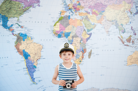 happy smiling little boy in captain cap and striped shirt with camera near world mapの写真素材