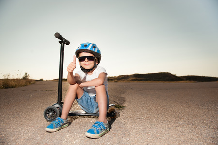 cute smiling kid with thumb up finger wearing sunglasses and helmet sitting on scooter on road in sunset summerの写真素材
