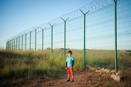 one little dirty caucasian boy refugee in empty desert field near long fencing with barbed wireの写真素材