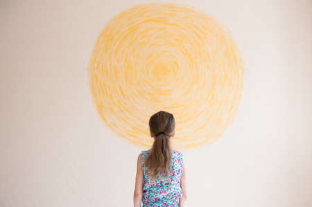 little caucasian girl with long hair looking at wall with painted yellow sun conceptの写真素材