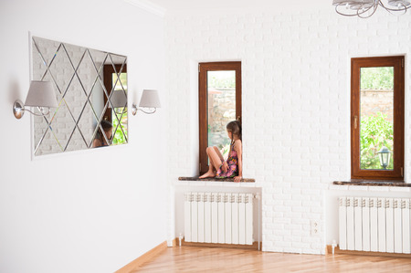lonely little girl sitting on window watching in to window on yardの写真素材