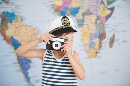cute small boy in captain hat taking picture with retro film camera on map backdropの写真素材