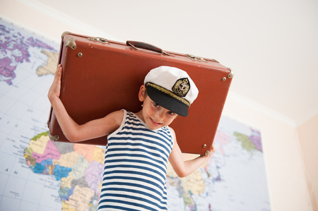 strong kid in captain cap holding heavy suitcase on his back on world map backgroundの写真素材
