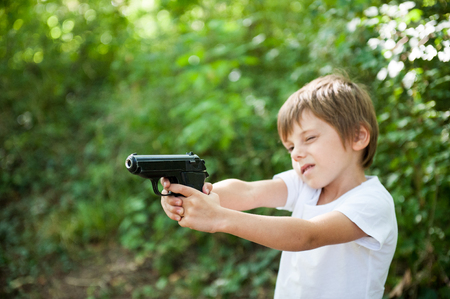 cute little boy in white shirt taking aim with black gun in green forest copyspaceの写真素材
