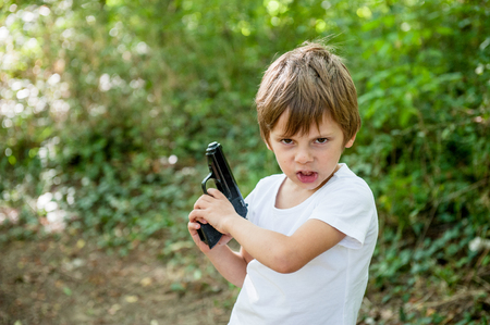 angry small caucasian boy with grimace on his face holding real black gun outdoorsの写真素材