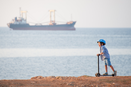 funny little caucasian boy in helmet shouting standing on scooter near sea with big shipの写真素材