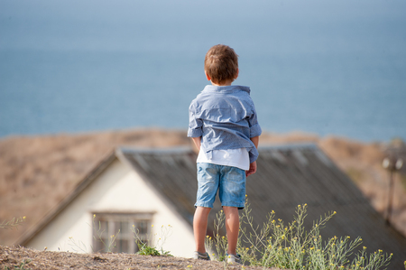 lonely small caucasian boy in shirt and shorts on old house and ocean shore backdropの写真素材