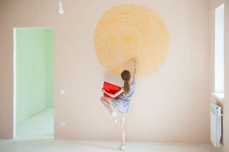 slim little caucasian girl with red palette brush painting yellow sun on wall in new roomの写真素材