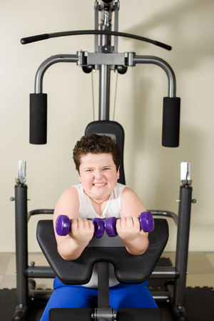 strong fat caucasian boy lifts dumbbells with hard effort on exercise machine in gymの写真素材