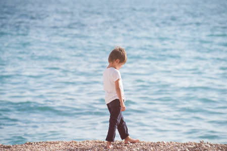 pensive sad caucasian lonely small boy walking along sea coast beach in autumn seasonの写真素材