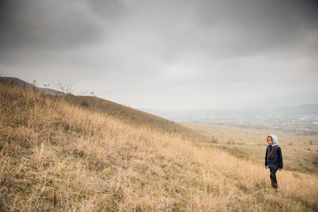 one lonely caucasian small girl in jacket with hood in overcast mountain on slope