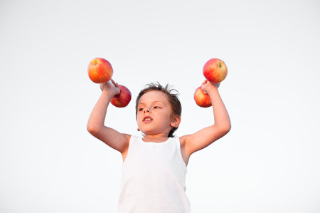 strong healthy cute caucasian kid in white shirt lifting dumbbells made from fresh applesの写真素材