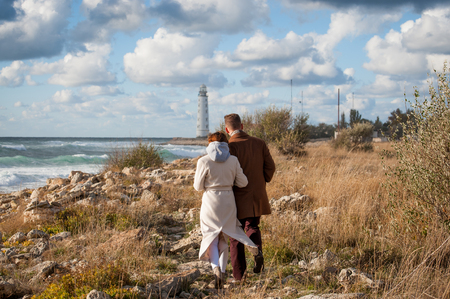 young pair male and woman in coat walking along stormy sea coast towards lighthouseの写真素材