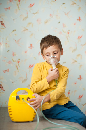 cute little caucasian boy in yellow shirt making inhalation at home using electric medical gearの写真素材