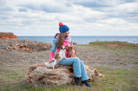 beautiful young woman in hat and track suit together with two little chihuahua dogs on sea shore in cold weather spring autumn dayの写真素材