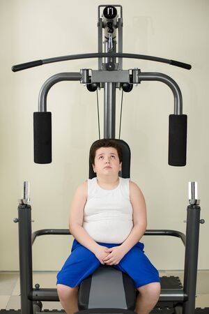fat thick little boy in white shirt sitting on exercise sport machine indoorsの写真素材
