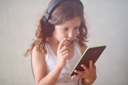 pensive caucasian little girl in earpieces holding black tablet pad