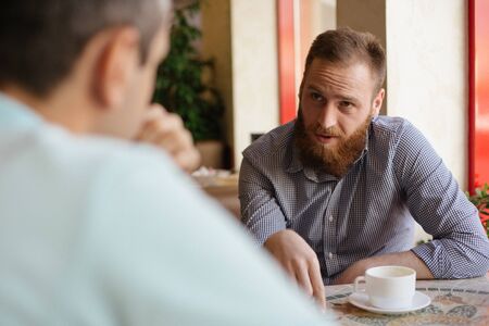 young bearded man explain something to his interlocutor friend sitting indoors cafe with cup of coffeeの写真素材