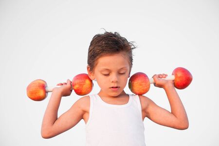 strong little caucasian boy in white tank top lifting dumbbells of apple during training sport workout with mind concentrationの写真素材