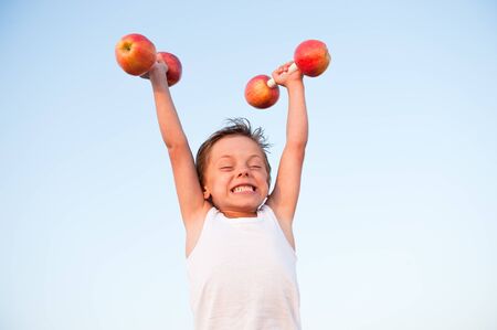 strong little caucasian boy in white tank top lifting dumbbells of apple during training workoutの写真素材