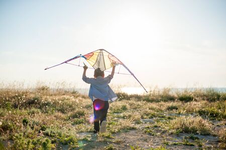 brave new world concept of little active boy running with kite above his head towards sunset sea horizonの写真素材