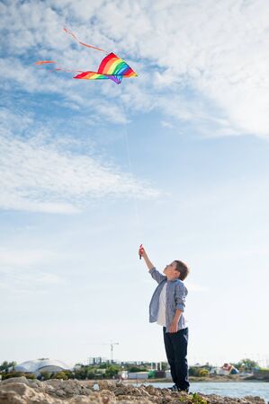 active little caucasian boy with flying multicolored kite on blue sky and sea shore rock backgroundの写真素材