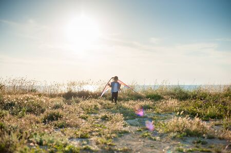free small kid with kite toy behind shoulders standing on sunset sea landscape with lens flareの写真素材
