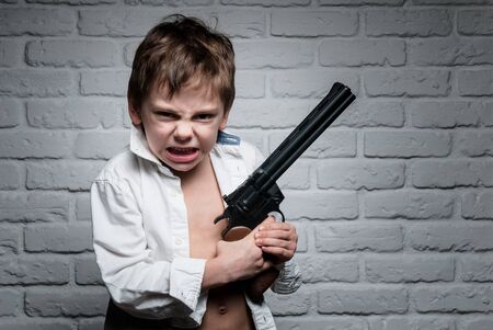 angry little boy in white shirt holding long revolver gun on brick wall backdropの写真素材