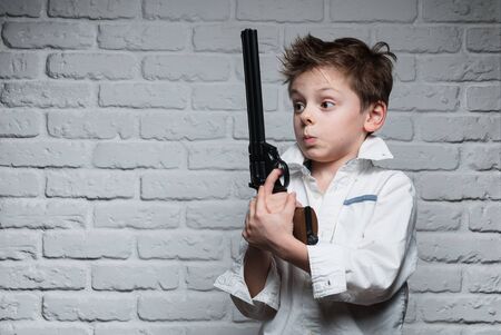 funny cute small kid holding big gun watching at it on brick wall background with copy spaceの写真素材