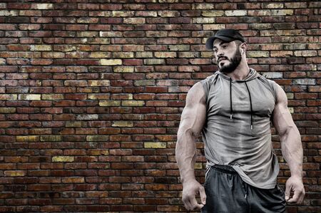 Powerful strong young bearded muscle man in sport hoodie shirt wearing cap and wireless headset with brick masonry old wall with copy spaceの写真素材