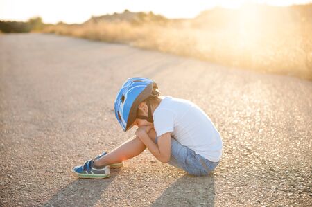 Little child in blue helmet sitting on asphalt road with closed eyes having leaned on knees on summer sunsetの写真素材